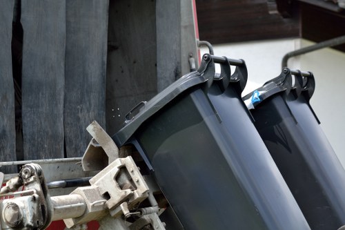 The image shows two black plastic wheelie bins secured to a metal frame on the back of a vehicle, likely a waste collection or rubbish removal truck. The bins are positioned side by side, with their lids closed, highlighting their smooth, durable plastic surface and slightly rounded edges. The metal attachment mechanism, which appears to be made of galvanized steel, connects the bins to the vehicle's framework, allowing for lifting and emptying. In the background, a wooden panel or fence is visible, along with parts of the vehicle's structure, suggesting an outdoor setting such as a driveway or a waste collection area near a building. The lighting suggests daytime conditions with natural light illuminating the scene, emphasizing the textures of the plastic and metal components. This setup aligns with a professional rubbish removal service operating in the vicinity of Covent Garden, London, and reflects typical equipment used by Office Clearance Covent Garden for waste collection and clearance tasks.