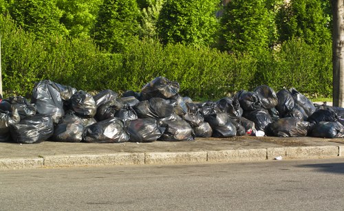 A long row of black rubbish bags, filled and tied at the top, are stacked along the edge of a paved street or driveway, with a curb separating them from the asphalt road. The bags appear to contain various types of waste materials, and are placed outdoors during daylight. Behind the line of rubbish bags, there is a dense, green hedge and tall trees with fresh, bright foliage, indicating a natural environment near a residential or park area in or near Covent Garden. The scene is clean and orderly in appearance, reflecting the type of waste removal service provided by Office Clearance Covent Garden, demonstrating a typical outdoor rubbish clearance arrangement in an urban or suburban setting.