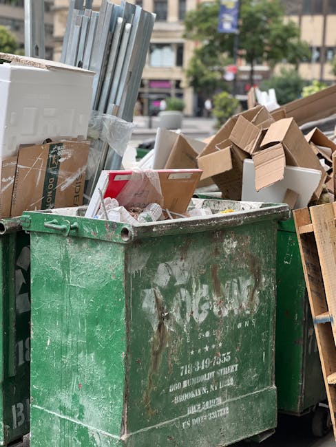 A large green wheeled waste bin filled with multiple red and black plastic bags containing bulky rubbish, positioned outdoors against a dark blue brick wall. The bags appear to contain construction or