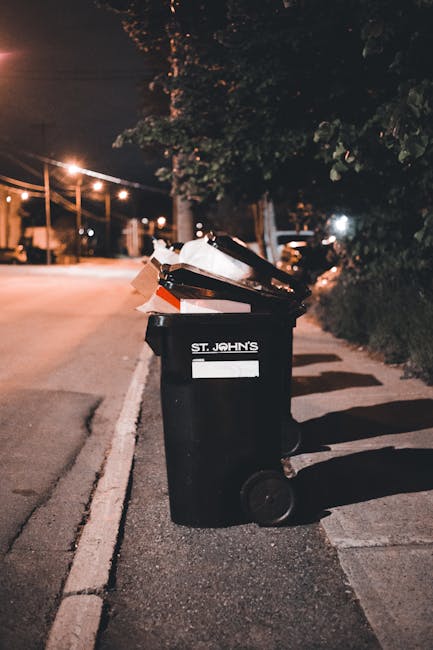 A narrow urban alleyway filled with various waste materials, including a large, weathered black plastic sack in the foreground, which appears to be partially open with contents not visible. Behind it,